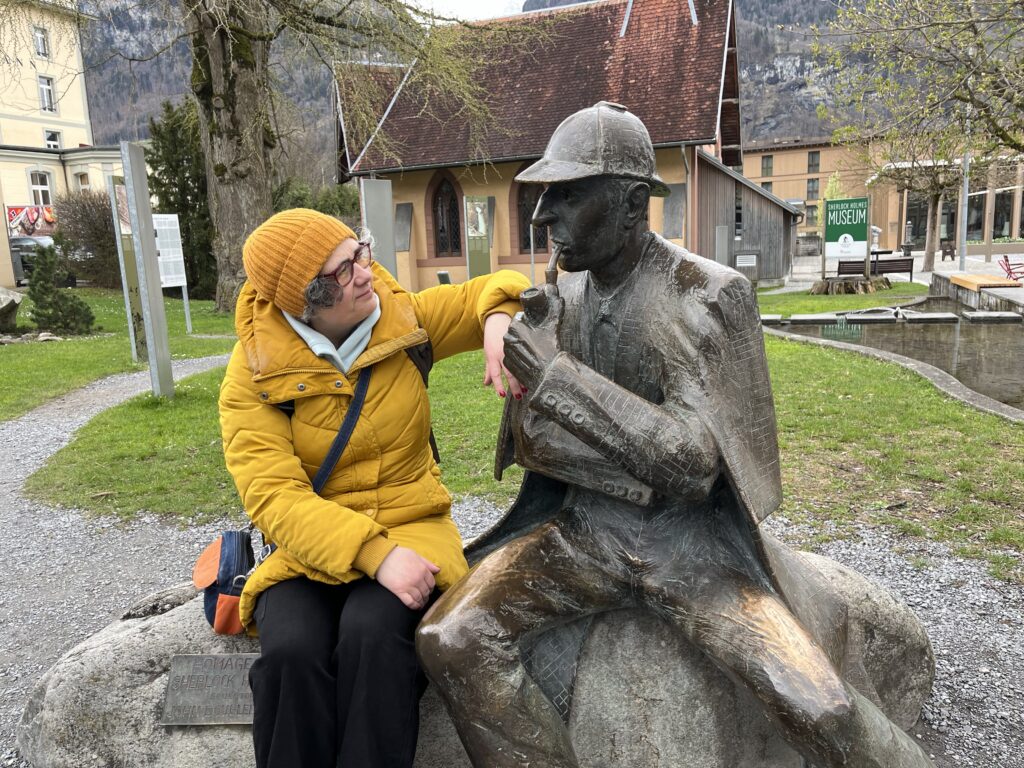 A photograph of a woman on the left in a yellow coat and hat seated and with her left arm resting on a bronze statue of Sherlock Holmes outside the Sherlock Holmes Museum in Meiringen, Switzerland